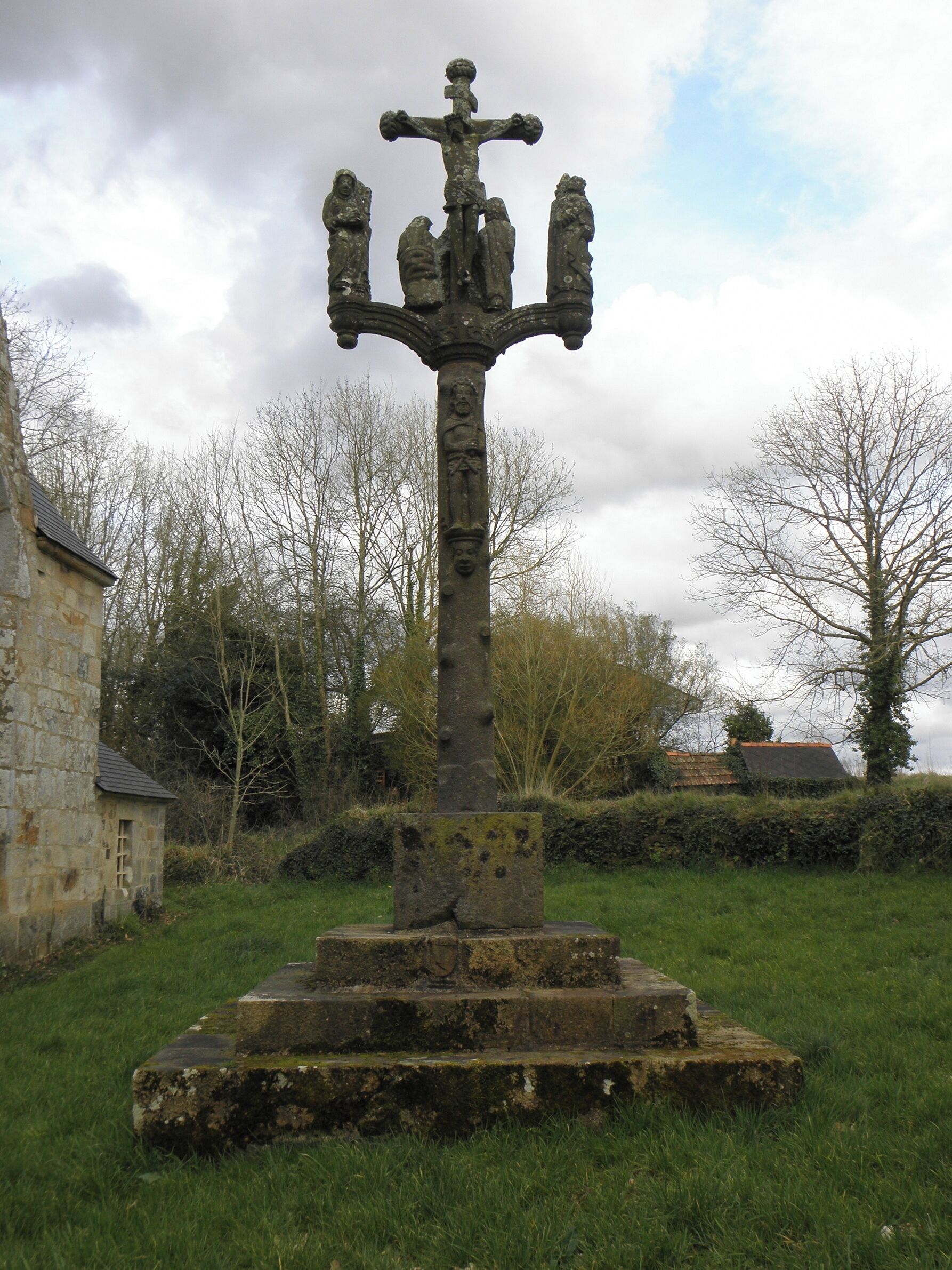 A stone cross with a crucifixion scene, surrounded by grass and trees.