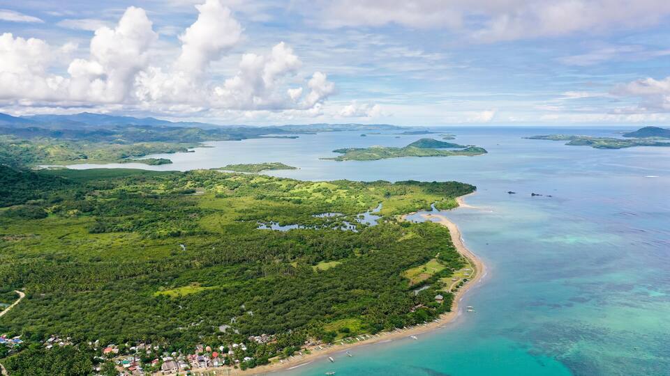 An aerial view of a coastal area with a beach, dense greenery, and several small islands in the distance.