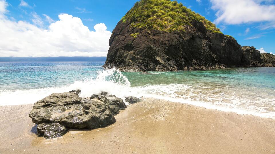 A beach with clear turquoise water, a large rock formation, and a small hill with greenery.