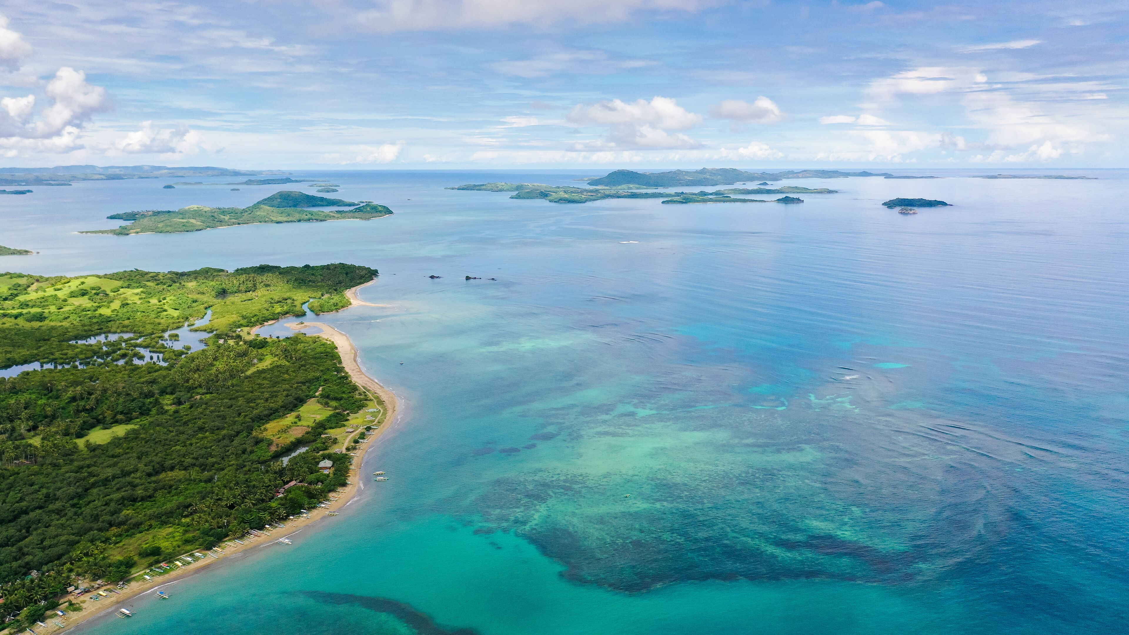 An aerial view of a coastal area with a beach, clear turquoise waters, and several small islands in the distance.
