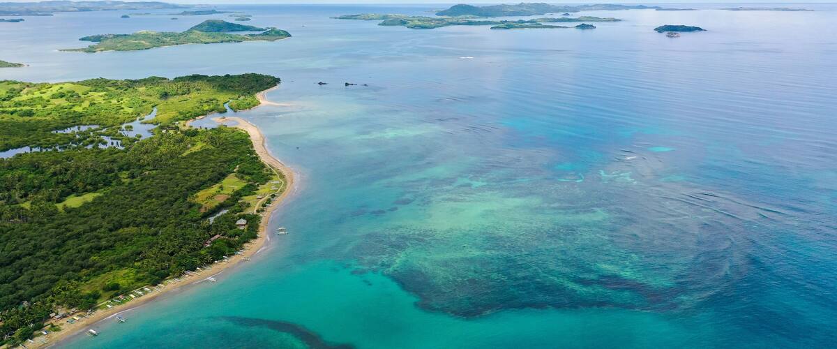 An aerial view of a coastal area with a beach, clear turquoise waters, and several small islands in the distance.