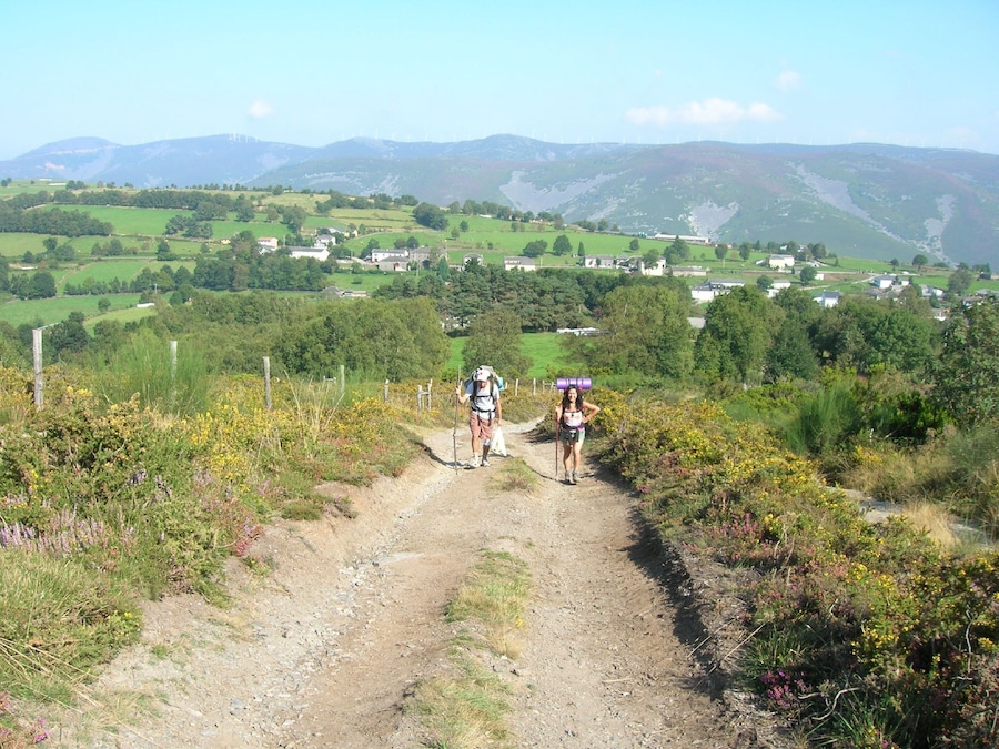 Un sendero de tierra con dos excursionistas, rodeado de campos verdes y casas a lo lejos.