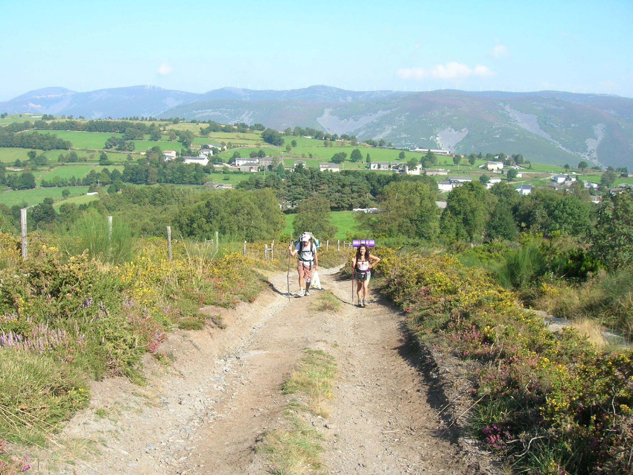 A dirt path with two hikers, surrounded by green fields and distant houses.