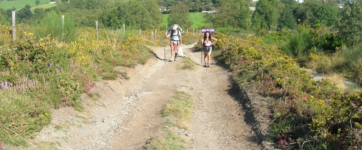 A dirt path with two hikers, surrounded by green fields and distant houses.