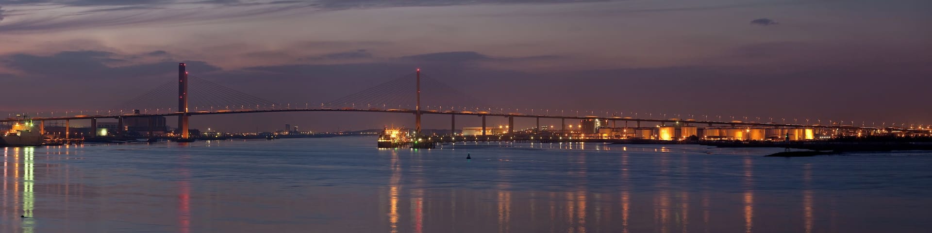 A large bridge spanning a body of water at dusk with city lights reflecting on the surface.
