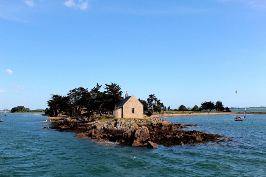 Pequeño edificio de piedra en una isla rocosa rodeada de agua tranquila y árboles.
