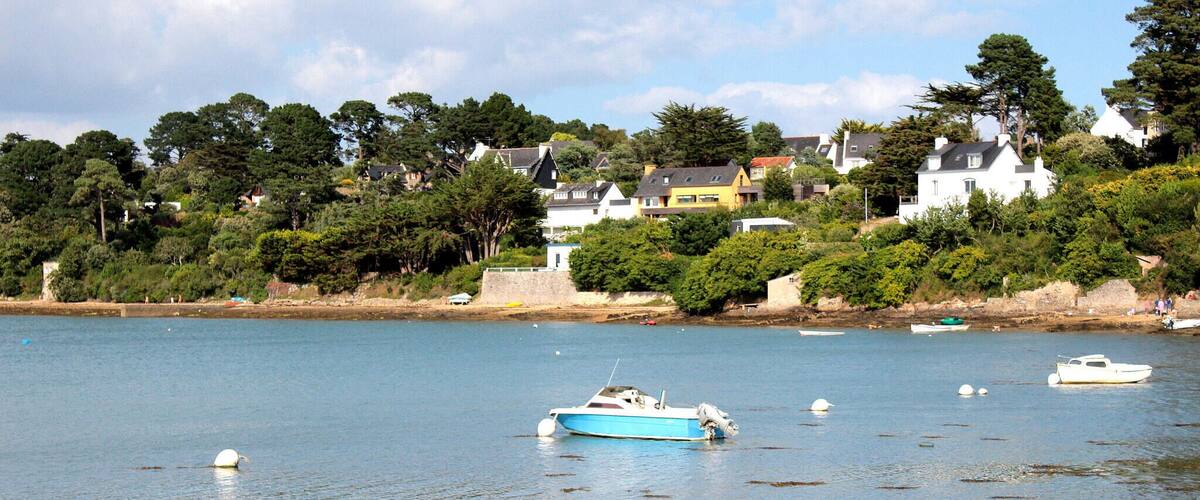 A coastal scene with a blue boat anchored in shallow water, surrounded by seaweed. In the background, there are houses on a hillside with trees and a clear sky.