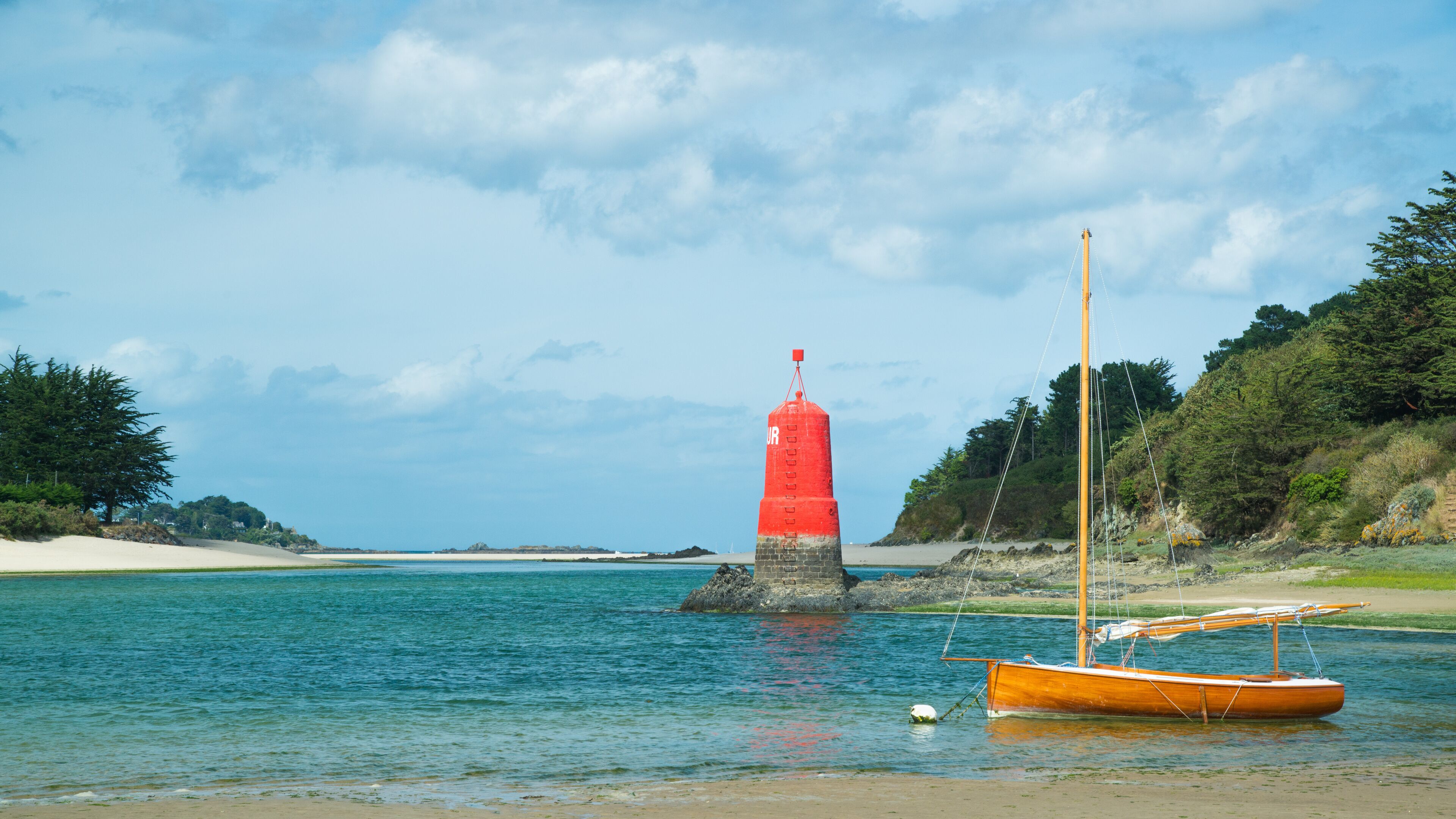 A red lighthouse on a rocky outcrop, a sandy beach, and a small orange boat anchored near the shore.