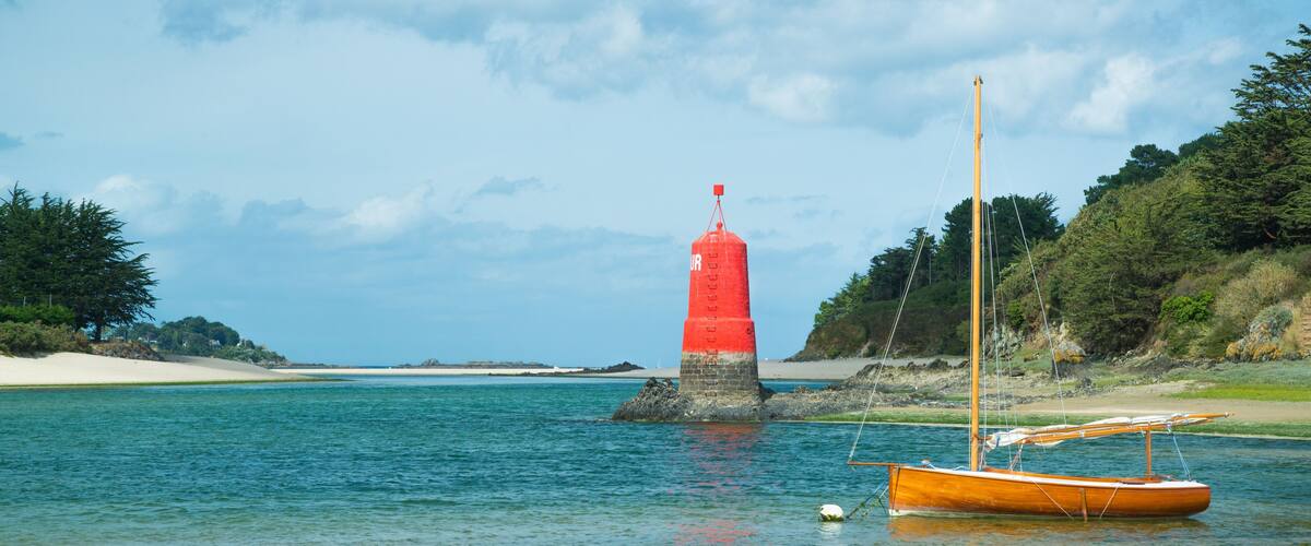 A red lighthouse on a rocky outcrop, a sandy beach, and a small orange boat anchored near the shore.