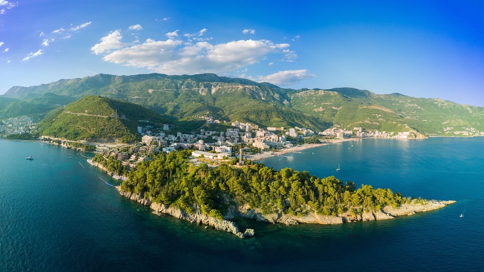 A coastal town with a mountainous backdrop, a small island with dense greenery, and clear blue waters.