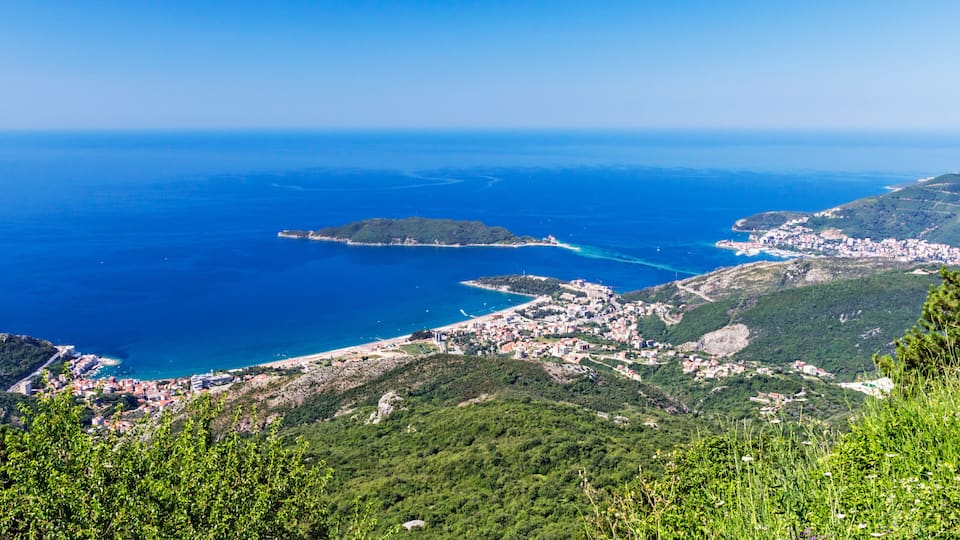 A coastal town with a clear view of the sea, a small island, and surrounding green hills.