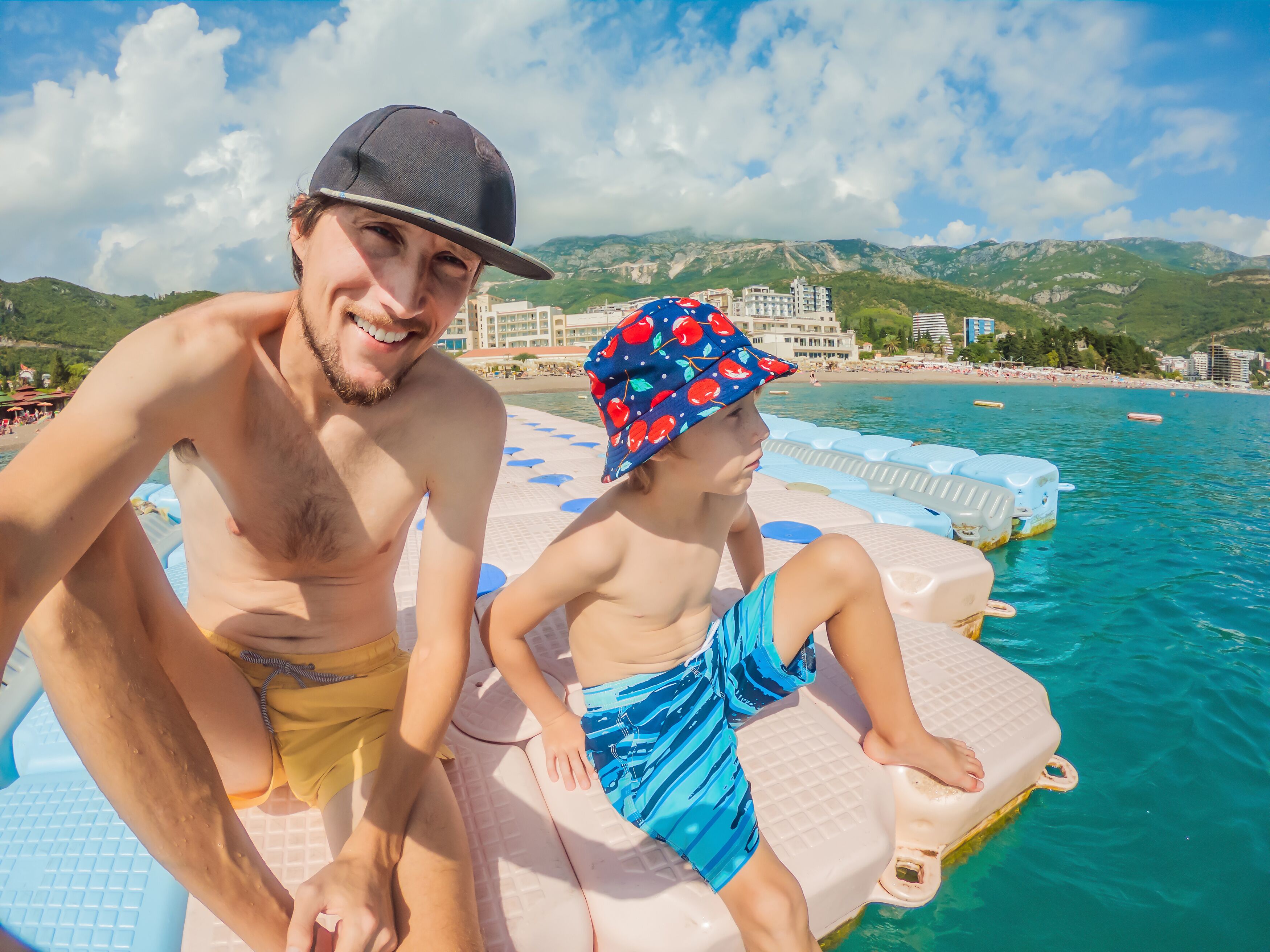 Un hombre y un niño en una plataforma flotante junto a una playa, con montañas al fondo.