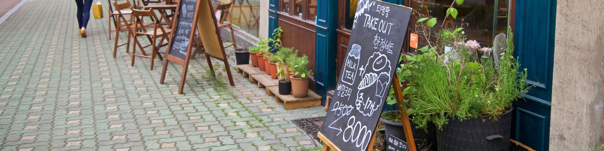 A street with outdoor seating, potted plants, and a chalkboard sign advertising tea and milk prices.