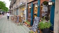 A street with outdoor seating, potted plants, and a chalkboard sign advertising tea and milk prices.