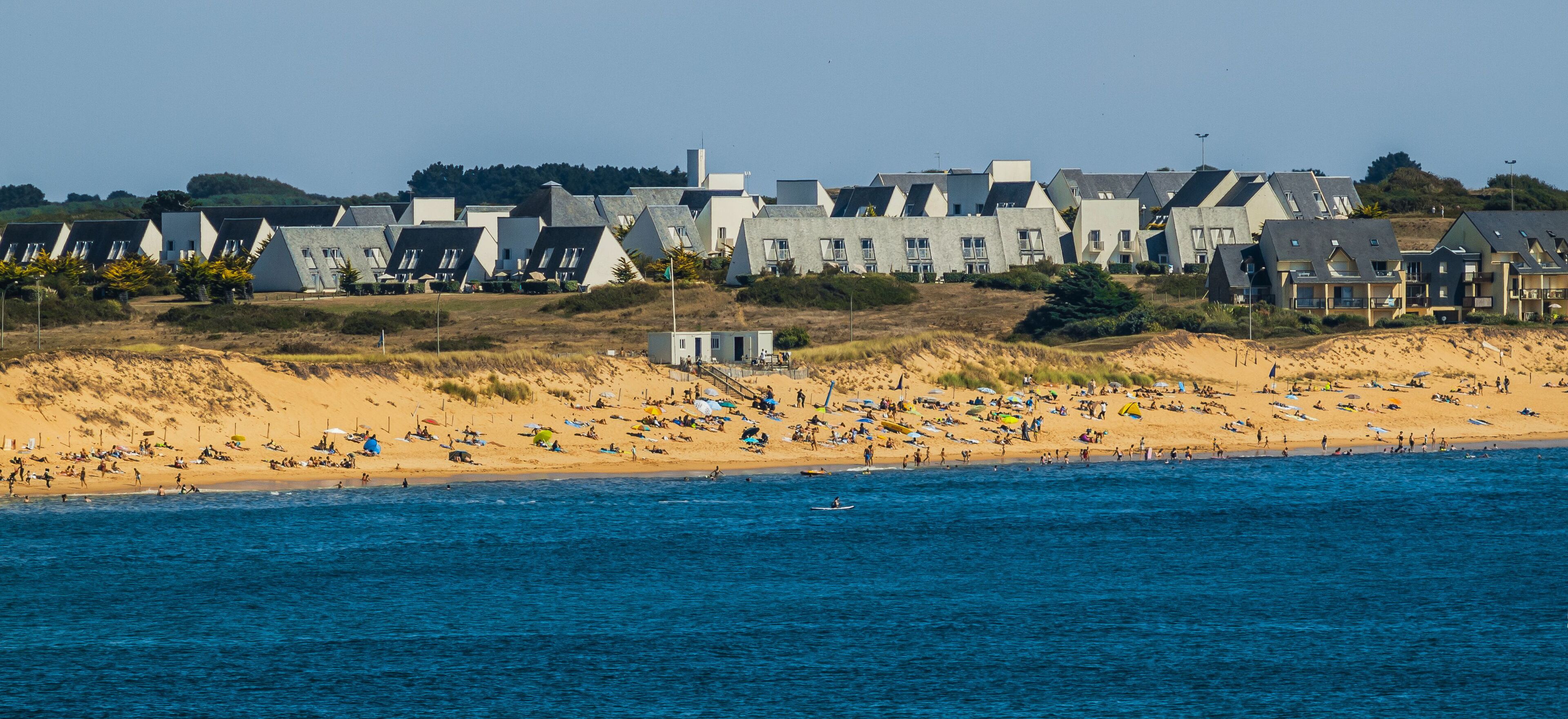 A beach with people sunbathing and swimming, and houses in the background.