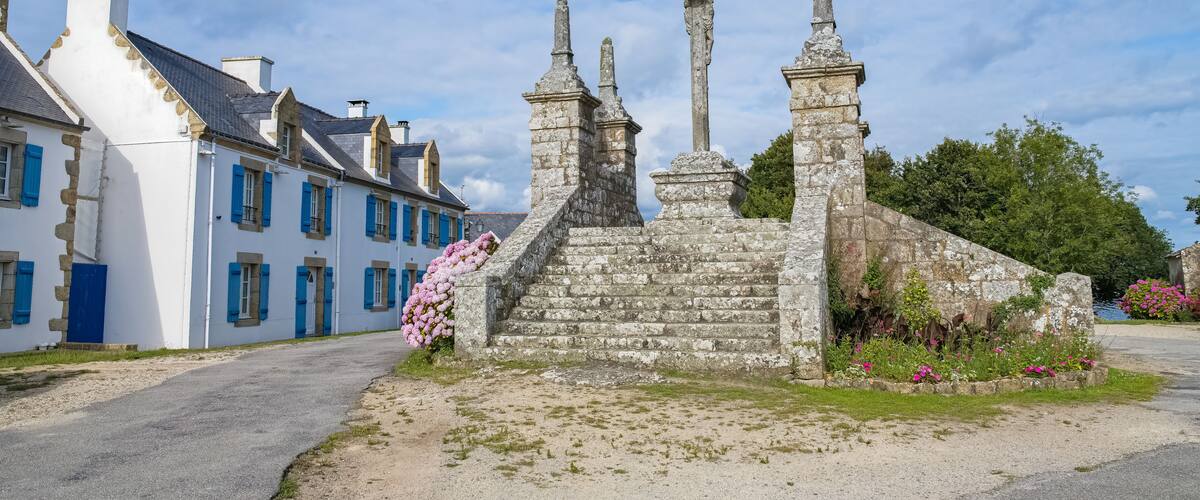 A stone monument with steps and statues, surrounded by a garden with pink flowers, and a white building with blue shutters.