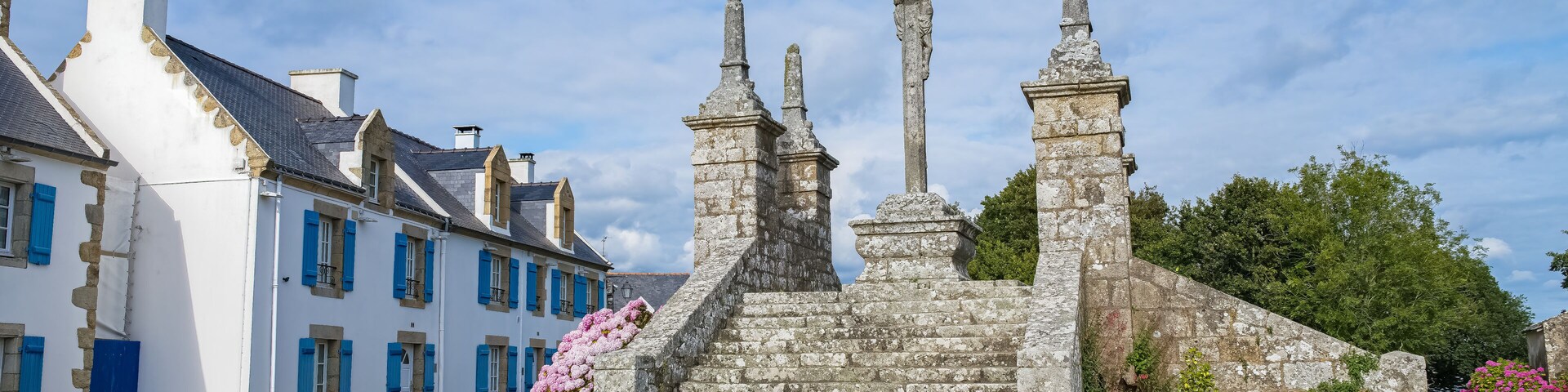 A stone monument with steps and statues, surrounded by a garden with pink flowers, and a white building with blue shutters.