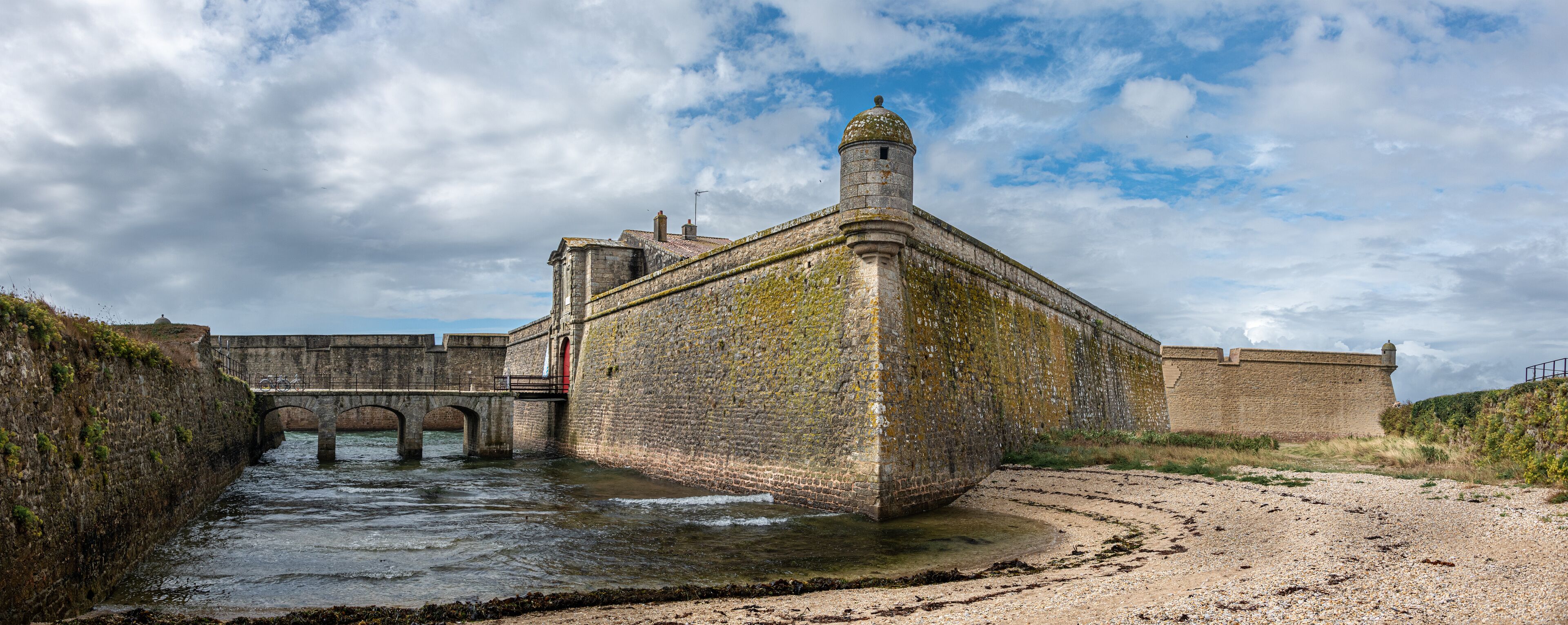 A historic stone fortification with a bridge over a river, surrounded by a gravel path and greenery.