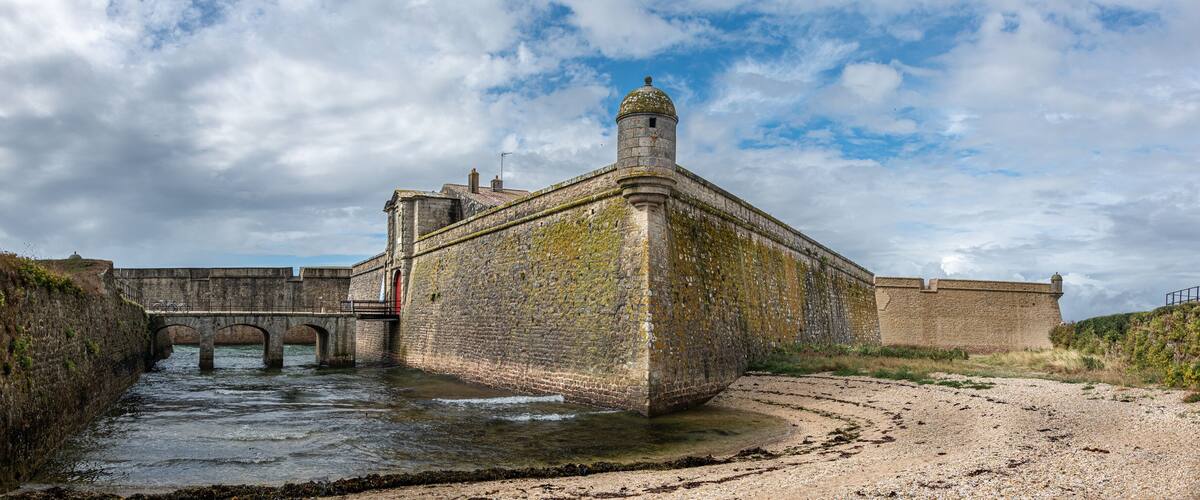 A historic stone fortification with a bridge over a river, surrounded by a gravel path and greenery.