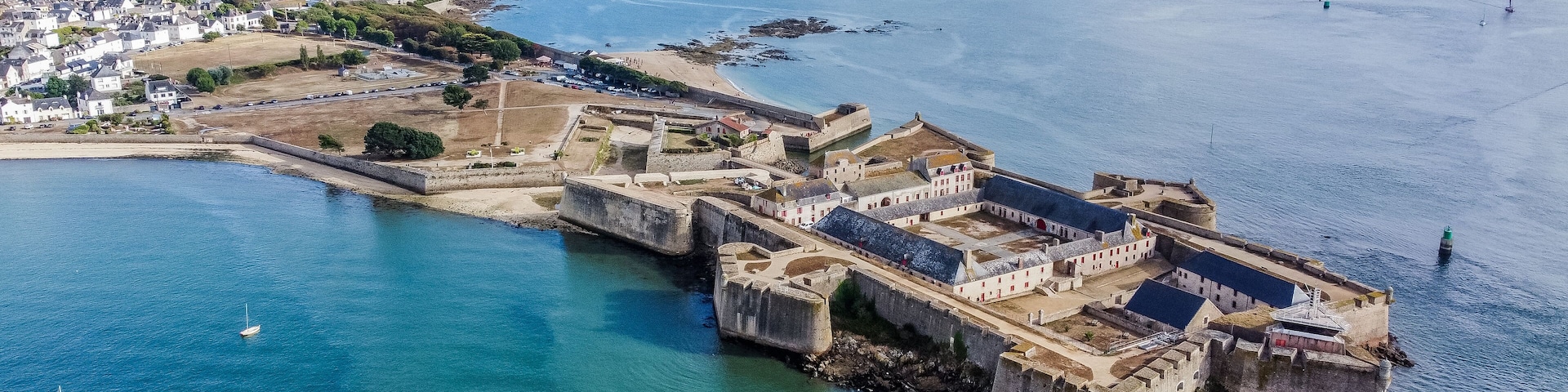 Una fortificación costera con un puerto, rodeada por un pueblo y el mar.