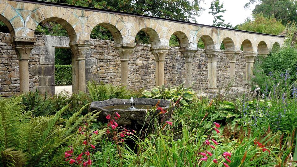 A stone bridge with arches, surrounded by lush greenery and a small pond.