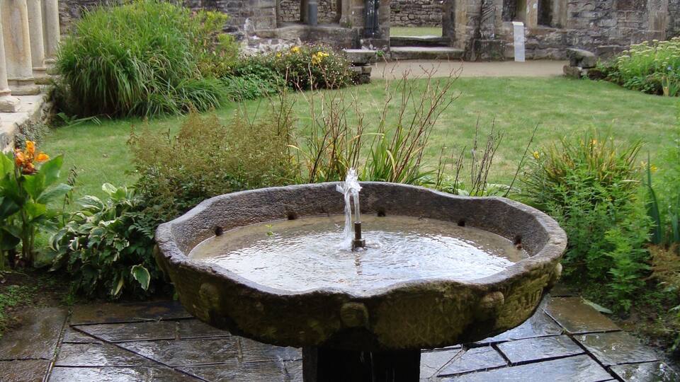 A stone fountain in a garden with a stone wall and arches in the background.