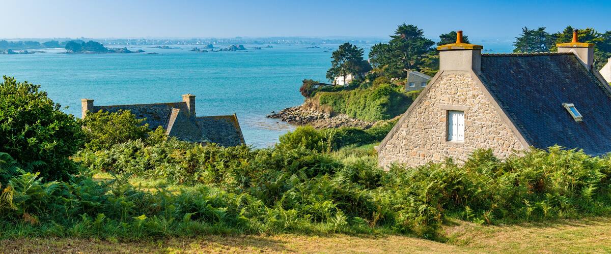 A coastal village with stone houses, lush greenery, and a view of the sea and distant islands.