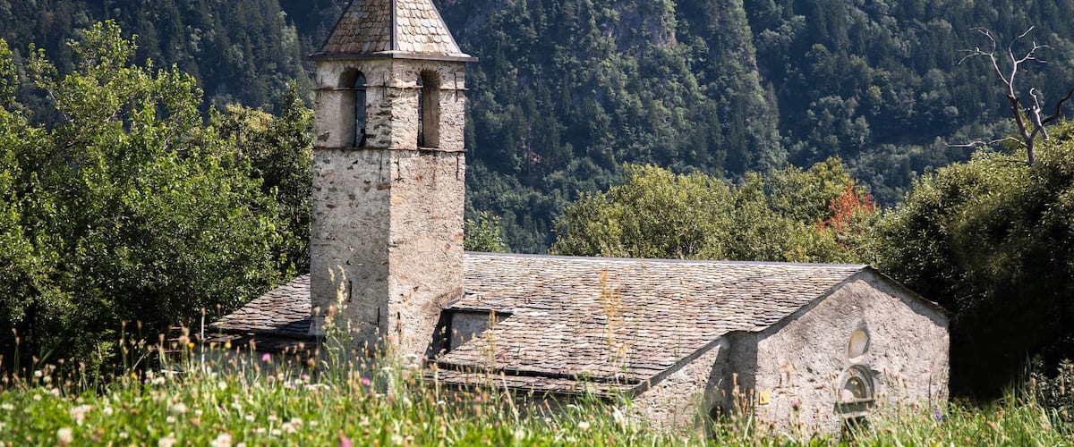 A stone church with a bell tower surrounded by lush greenery and a forested mountain backdrop.