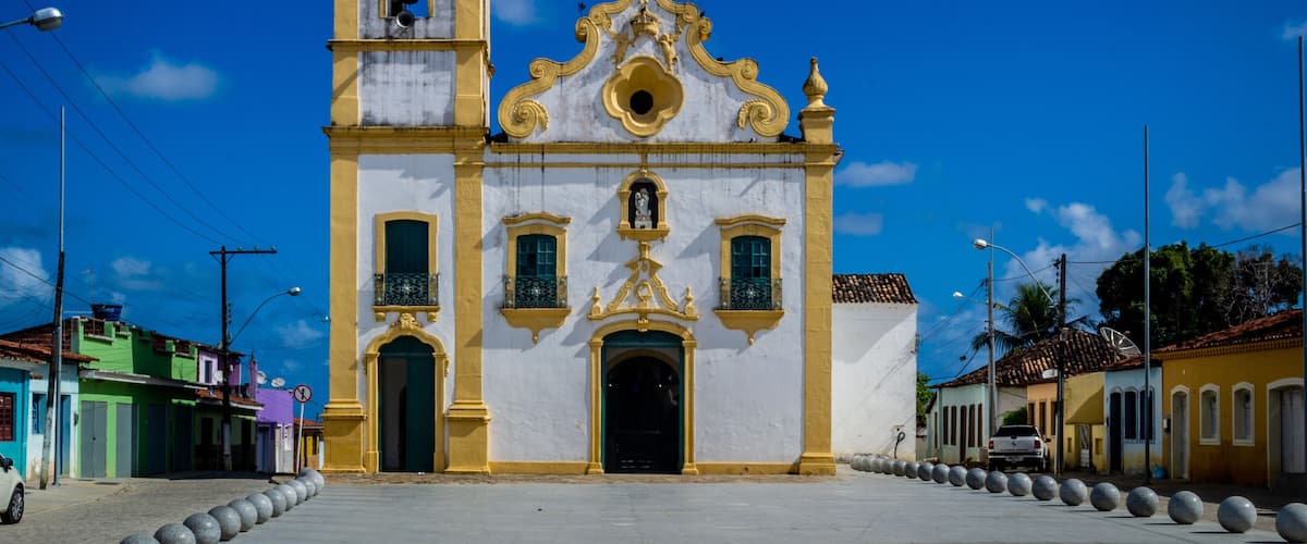 A white and yellow church with a bell tower and a cross on top, surrounded by a clear blue sky.