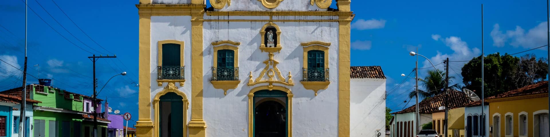 A white and yellow church with a bell tower and a cross on top, surrounded by a clear blue sky.