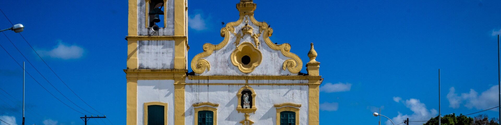 Una iglesia blanca y amarilla con un campanario y una cruz en la parte superior, rodeada por un cielo azul despejado.
