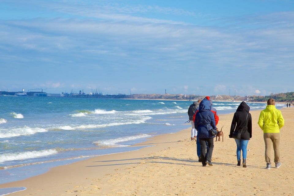 A beach with people walking along the shore, waves crashing onto the sand, and a distant industrial area with cranes and buildings.