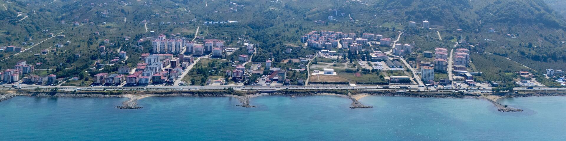 Aerial drone view of Arsin, Trabzon coastline with residential buildings and green hills. Black Sea shore meets lush mountains, showing urban landscape, nature, and coastal development in Turkey.
