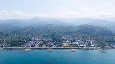Aerial drone view of Arsin, Trabzon coastline with residential buildings and green hills. Black Sea shore meets lush mountains, showing urban landscape, nature, and coastal development in Turkey.