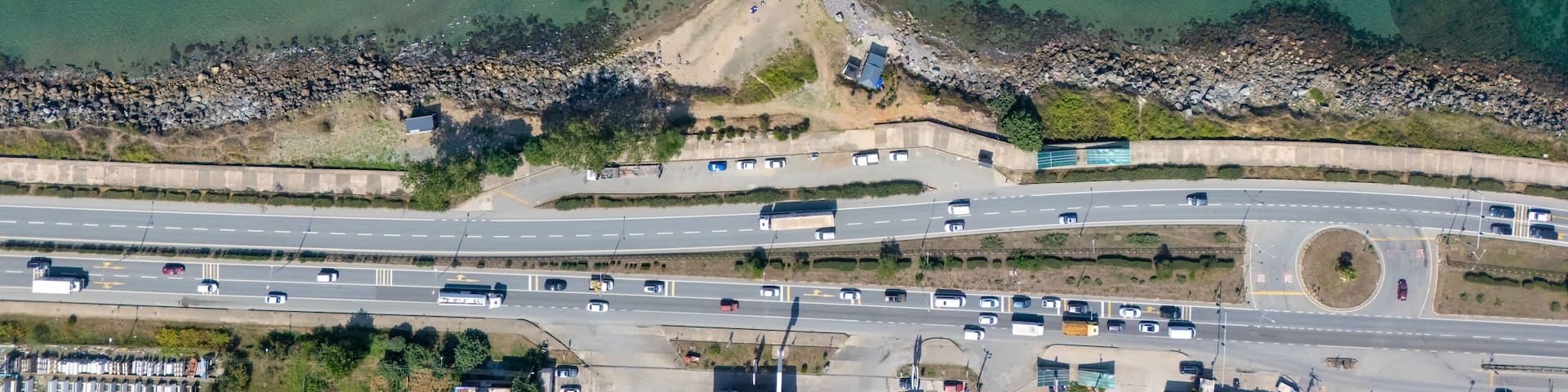 High-angle drone shot of a T-shaped breakwater in Arsin, Trabzon, Turkey, where the Black Sea meets the coast, with roads, buildings, and green-blue water blending urban and natural landscapes.