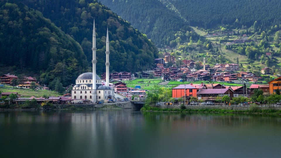 A mosque with two minarets, a lake, and a mountainous backdrop.