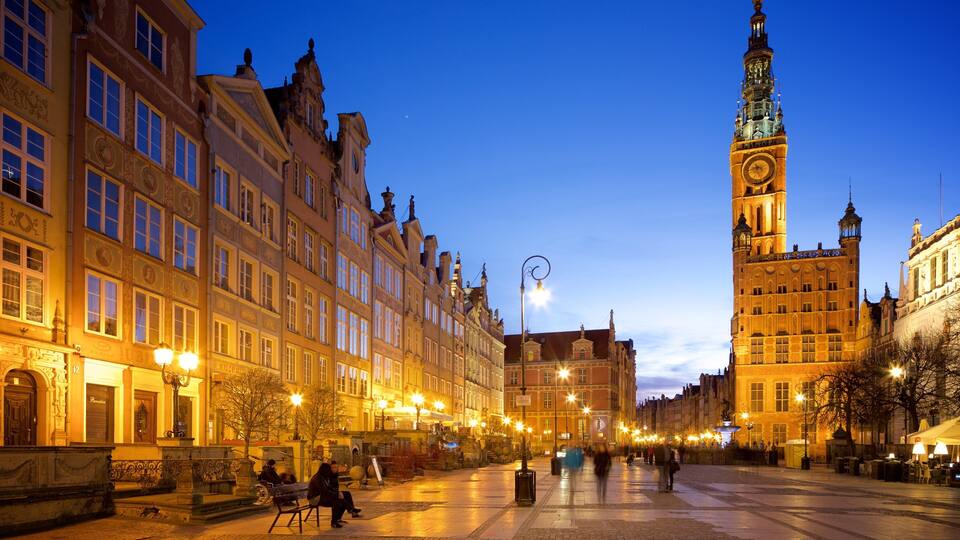 A historic European square with ornate buildings, a prominent clock tower, and a well-lit street at dusk.