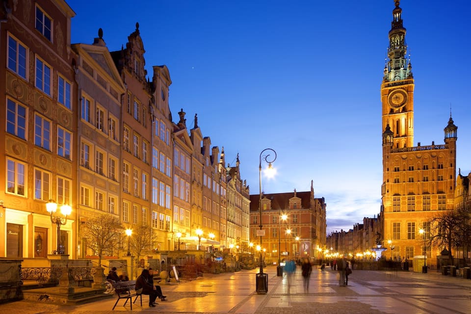 A historic European square with ornate buildings, a prominent clock tower, and a well-lit street at dusk.