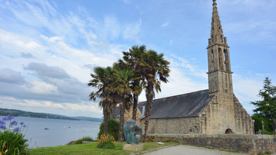 A historic stone church with a tall bell tower by a lake, palm trees, and a sculpture.