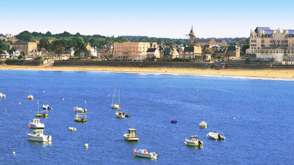 A coastal town with a sandy beach, numerous boats in the water, and historic buildings along the shore.