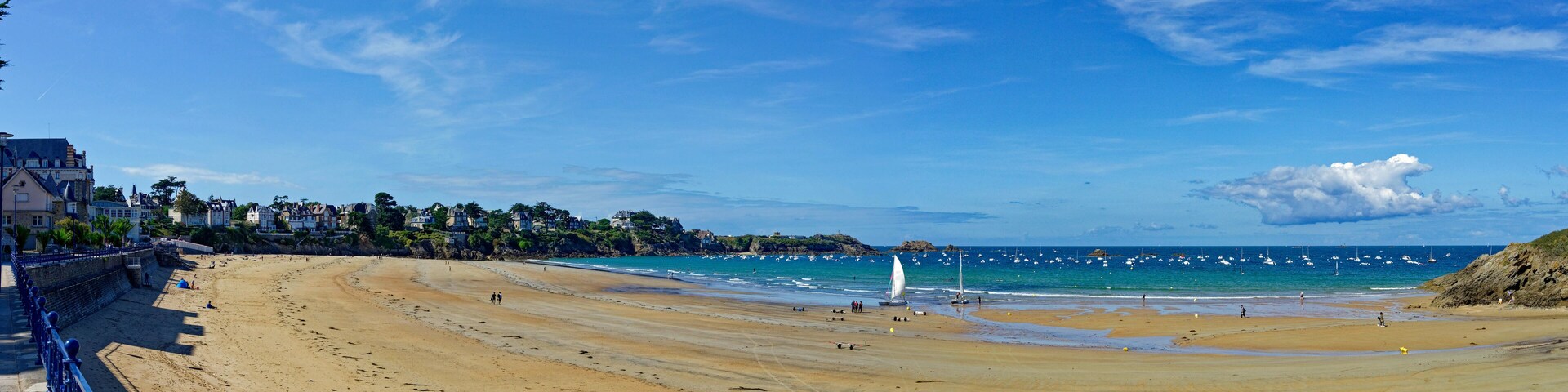 Uma extensa praia de areia com céu azul límpido, um veleiro e um afloramento rochoso.