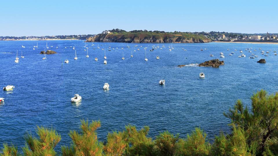 A coastal town with numerous boats on the water, rocky outcrops, and a clear blue sky.