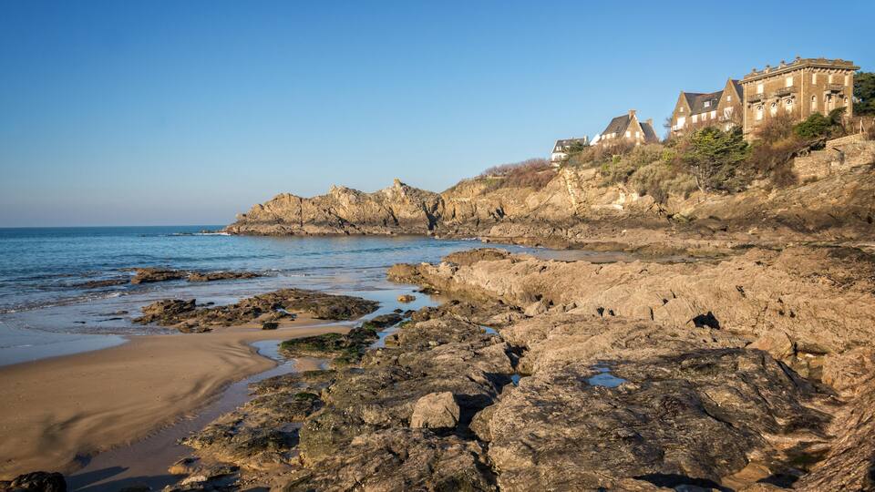 A rocky coastline with a clear blue sky, a sandy beach, and historic buildings on a hillside.