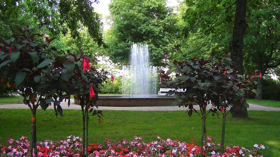 A park with a central fountain, surrounded by lush greenery and flowering plants.
