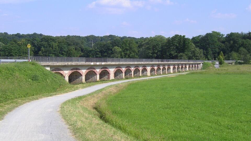 A stone bridge with multiple arches over a road, surrounded by green fields and trees.