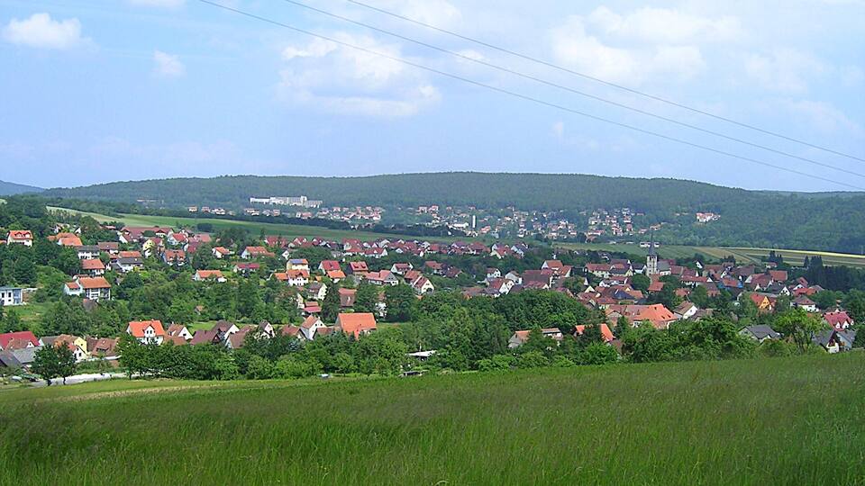 A town with red-roofed houses, green fields, and a hill in the background.