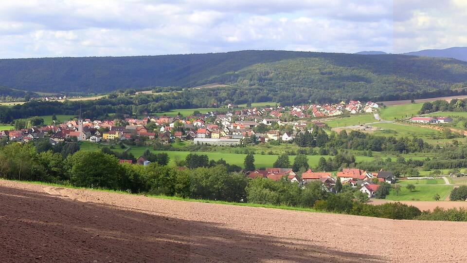 A rural village nestled in a valley with rolling hills and a river in the background.