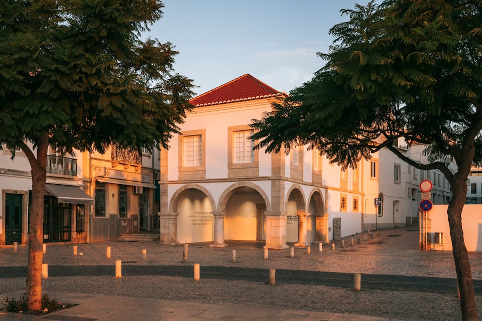 Un bâtiment historique, avec un toit rouge et des arcades, entouré d’un pavé et d’arbres.