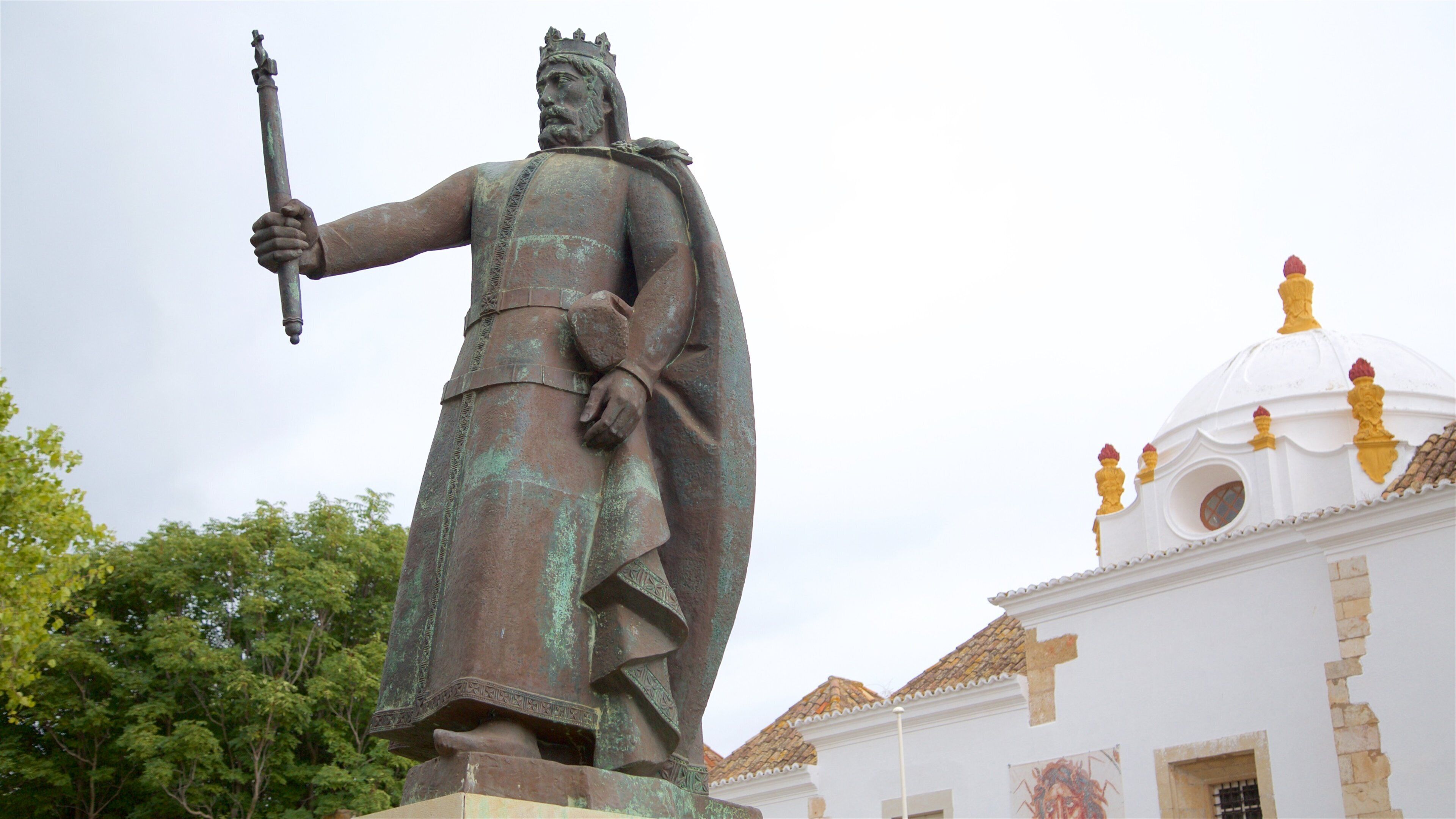 En statue af en figur med en stav, foran en hvid bygning med en kuppel og gyldne detaljer.