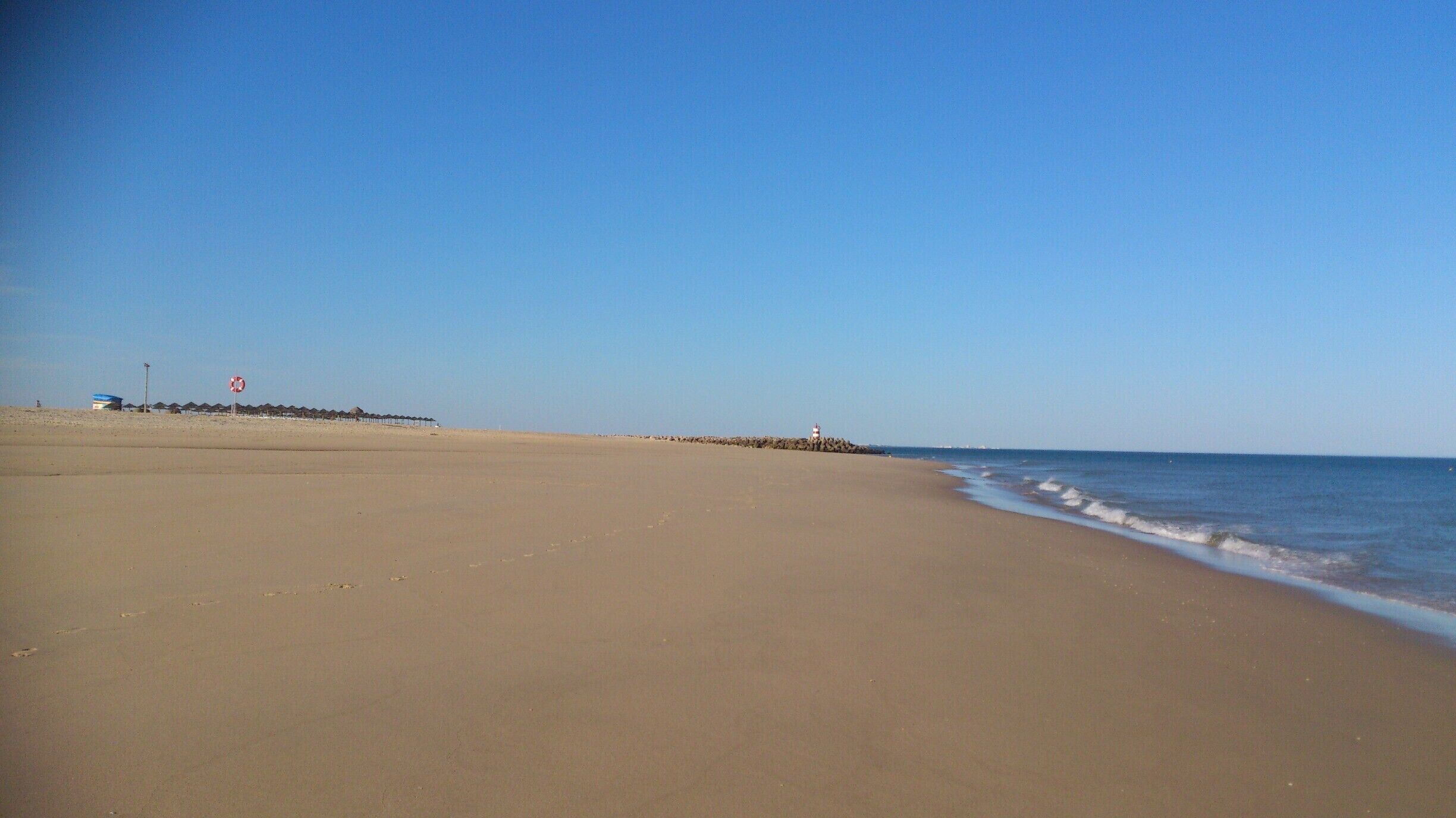 A wide sandy beach with gentle waves, a clear blue sky, and a lighthouse in the distance.